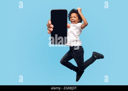 Voller Länge, lebhaft energisch kleinen Jungen springen vor Glück und zeigt Handy-Bildschirm und überrascht Sieger Gesicht, Kind fliegt, Gefühl in Stockfoto