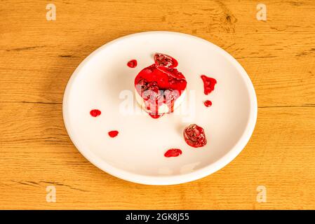 Tarta de queso individual con mucha mermelada de frambuesa sobre plato de porcelana blanca y mesa de madera Stockfoto