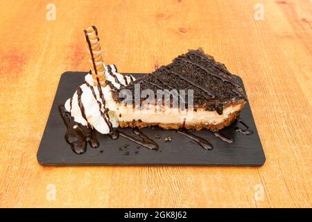 Köstlicher Käsekuchen mit Stücken von oreo, Schokoladensirup, Creme und einer Waffel auf einem schwarzen Schieferteller Stockfoto