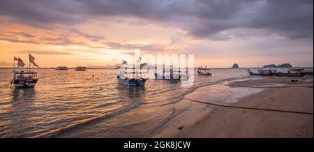 Weitwinkel mit malerischer Aussicht auf Holzboote auf dem plätschernden Meer, das die sandige Küste bei Sonnenuntergang in Malaysia unter bewölktem Himmel wäscht Stockfoto