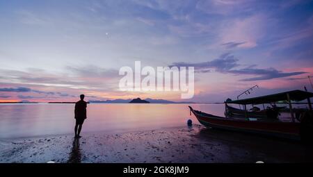 Ganzkörperrücken der Silhouette eines nicht erkennbaren Touristen, der an nassem Sandstrand in der Nähe des Bootes steht und das Meer unter dem Sonnenuntergang in Malaysia bewundert Stockfoto