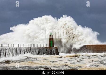 Riesige schäumende Meereswellen, die gegen steinerne Wellenbrecher krachen, mit altem Leuchtturm-Turm gegen den blau bewölkten Himmel im Hafen von Viavelez in Asturien, Spanien Stockfoto