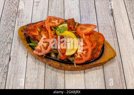 Tandoori-Hähnchen auf einem heißen Teller mit Tomaten, Pfeffer, Zitrone und Koriander als Garnitur auf einem Holztisch gehackt Stockfoto