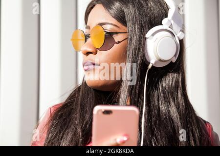 Trendige junge langhaarige afroamerikanische Frau in roter Jacke und Sonnenbrille, die Musik über Kopfhörer und Mobiltelefon hört, während sie su genießt Stockfoto