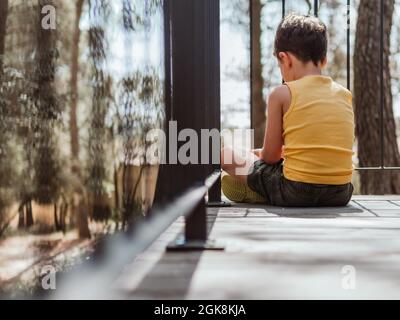 Rückansicht des einsamen beleidigten Kindes, das an sonnigen Tagen auf der Holzterrasse des Hauses im Wald sitzt Stockfoto