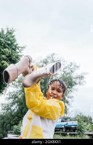 Lächelndes, ethnisches Kind in Slicker, das bei regnerischem Wetter aus Gummistiefeln Wasser gießt, während es die Kamera anschaut Stockfoto