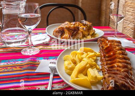 Rippchen mit Barbecue-Sauce und Kartoffeln, serviert in einem peruanischen Restaurant mit einem Krug Wasser und einem Brattopf auf einer bunten Tischdecke Stockfoto