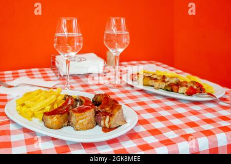 Gebratene Schweineribs mit Barbecue-Sauce und Kartoffeln auf einem Restauranttisch mit einem Set für zwei Personen, die Wasser trinken Stockfoto