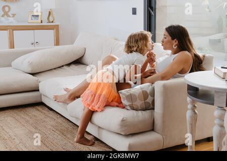 Seitenansicht einer wertigen Frau und eines Mädchens mit poulierenden Lippen, die die Hände halten, während sie sich auf der Couch zu Hause angucken Stockfoto