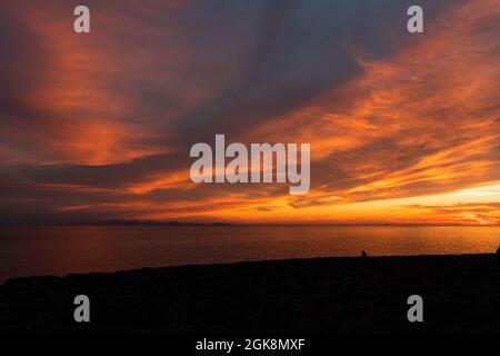 Spektakulärer Blick auf die Silhouette des Reisenden, der beim Sonnenuntergang den Ozean vom Strand aus unter einem farbenprächtigen, wolkigen Himmel betrachtet Stockfoto