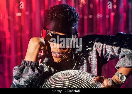 Trendige ethnische Frau in Cyberpunk-Brille und Armbanduhr berühren Disco-Ball in Lichtstrahl Stockfoto