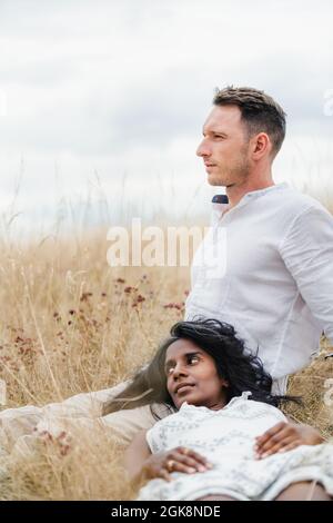 Verträumte indische Frau, die auf dem Oberschenkel eines männlichen Geliebten liegt, während sie im Herbst auf der Wiese unter dem wolkigen Himmel wegschaut Stockfoto