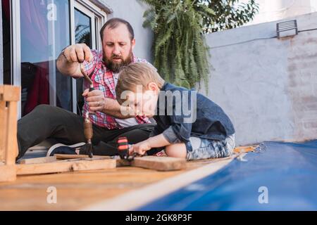 Bärtiger Vater unterrichtete Sohn mit Hammer, der am Wochenende auf der Promenade mit Holz arbeitete Stockfoto