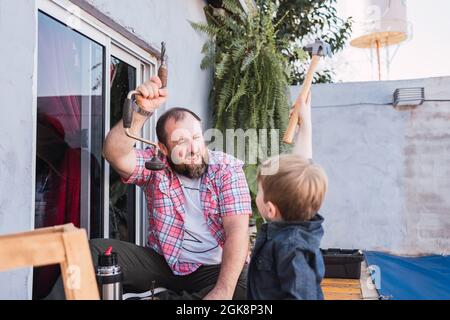 Bärtig fröhlicher Vater unterrichtet Sohn mit Hammer arbeiten mit Holz, während auf der Promenade am Wochenende sitzen Stockfoto