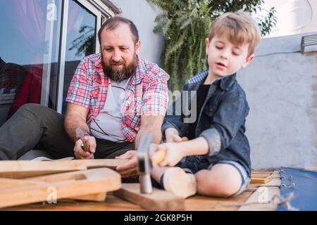 Bärtiger Vater unterrichtete Sohn mit Hammer, der am Wochenende auf der Promenade mit Holz arbeitete Stockfoto
