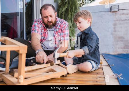 Bärtiger Vater unterrichtete Sohn mit Hammer, der am Wochenende auf der Promenade mit Holz arbeitete Stockfoto