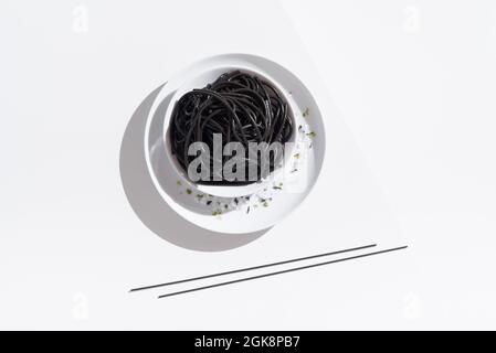 Von oben aus Keramik-Schüssel mit leckeren Spaghetti mit schwarzer Tintenfisch-Tinte mit Essstäbchen auf weißem Hintergrund Stockfoto