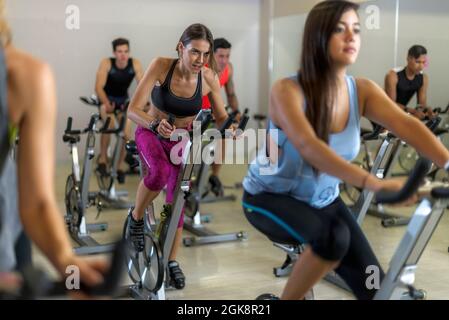 Fokussierte, schweißtreibende Sportlerin beim Radfahren in aktiven Trainingsbekleidung während des Gruppentrainings im geräumigen Fitnesscenter mit modernen Geräten Stockfoto
