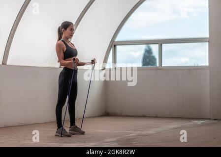 Entschlossene junge ethnische Athletin in Sportbekleidung, die mit einem Gummiband ausarbeitet und dabei Bizeps macht, während sie nach vorne blickt Stockfoto