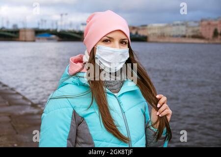 Eine junge Frau aus dem Kaukasus in warmen Kleidern und einer medizinischen Maske geht im Herbst an einem bewölkten Tag am Damm der Stadt entlang Stockfoto