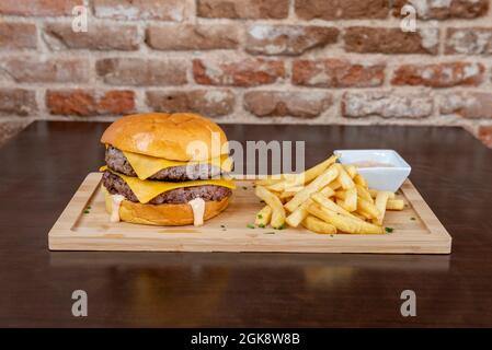 Double Burger mit geschmolzenem Cheddar-Käse und Mayonnaise-Sauce, garniert mit Pommes auf Holzbrett Stockfoto