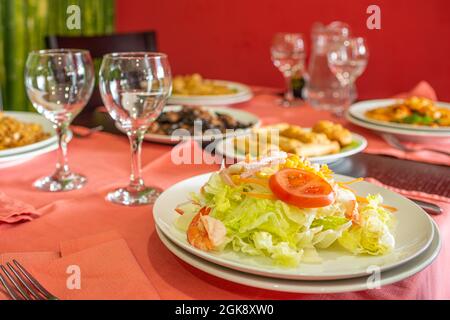 Eisbergsalat mit Tomaten, Mais, Tomatenscheiben, Schinken und Karotten mit gekochten Garnelen auf dem Tisch mit roten Tischdecken und Kristallgläsern Stockfoto