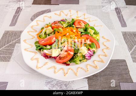 Gemüse- und Salatsalat mit Tomatenscheiben, geriebener Karotte, gewürfelter Gurke und weißem Teller auf grauer Tischdecke Stockfoto