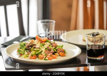 Caesar-Salat mit Kirschtomaten, gebratenem Speck und Gemüsesprossen. Glas mit Cola in einem Restaurant Stockfoto