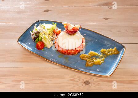 Burrata-Käse auf Tomatentartare, ein wenig Serrano-Schinken und grüner Salat auf einem blauen Tablett Stockfoto