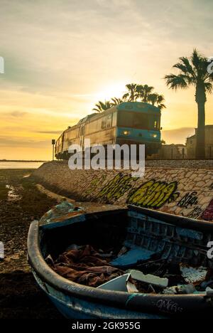Faro, Portugal - 12. April 2021: Altes Boot am Strand, Pastel-Sonnenuntergang in Faro als ein Zug vorbeifährt, Palmen und die Sonne dahinter Stockfoto