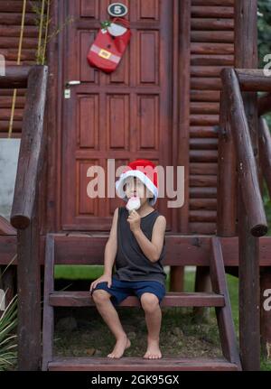 Lateinischer Junge, der zur Weihnachtszeit ein Eis in einem Weihnachtsmannhut genießt und auf der Treppe vor einer braunen Hütte sitzt. Vertikal, Sommer Stockfoto