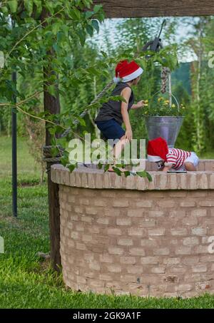 Zwei Kinder mit Weihnachtsmannmützen spielen auf einem geschlossenen Ziegelwasserbrunnen zur Weihnachtszeit. Grüne Vegetation im Hintergrund. Sommer. Vertikal Stockfoto