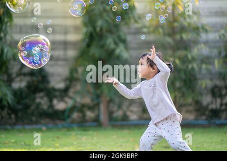 Flacher Fokus eines niedlichen südostasiatischen Kindes, das versucht, eine große Blase in der Luft im Garten zu berühren Stockfoto