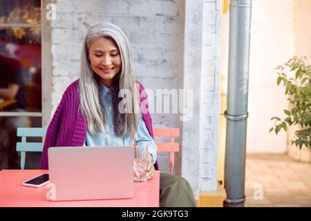 Positive ältere Frau mit Glas und Smartphone schaut auf den Laptop-Bildschirm auf der Terrasse des Cafés im Freien Stockfoto
