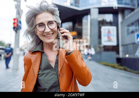 Glückliche, grauhaarige ältere Frau mit Brille spricht auf dem Handy auf der Stadtstraße Stockfoto