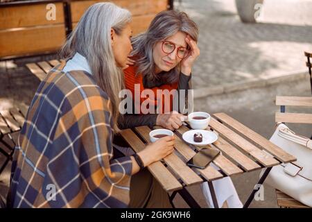 Deprimierte reife Dame mit Brille und vorsichtiger Freundin si im Straßencafé am Herbsttag Stockfoto