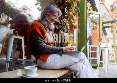 Positive ältere Frau mit Brille arbeitet auf dem Laptop auf der Bank in der Nähe des Cafefensters Stockfoto