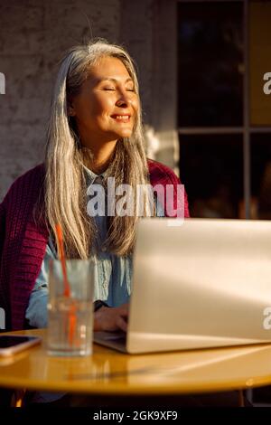 Fröhliche ältere asiatische Dame arbeitet am Laptop witting auf der Terrasse im Café bei Sonnenuntergang Stockfoto