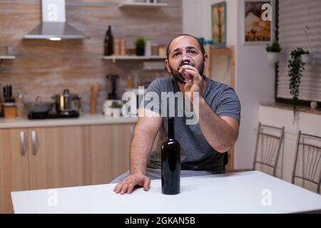 Nachdenklicher Mann, der ein Glas Wein trinkt, während er allein und deprimiert sitzt. Person mit Alkoholabhängigkeit mit einer Flasche alkoholisches Getränk, Schnaps, Alkohol, das Gefühl, heartbroken und verzweifelt Stockfoto