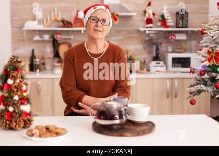 Porträt einer älteren Frau mit weihnachtsmann Hut am Tisch in Weihnachten dekoriert Küche genießen Wintersaison. Großmutter feiert weihnachtsfeiertage. Traditionelle hausgemachte Kekse auf dem Tisch Stockfoto