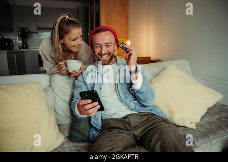 Kaukasisches Paar sitzt auf der Couch, trinkt Kaffee und bezahlt online mit einem Mobilfunkgerät Stockfoto