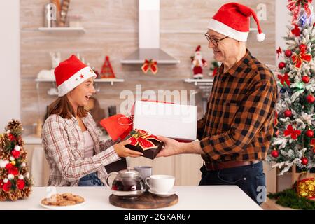 Großvater überraschende Enkelin mit Weihnachten Wrapper Geschenk genießen weihnachtsurlaub zusammen in dekorierter Küche. Kind feiert Wintersaison wechselnde Gegenwart mit Band darauf mit Opa Stockfoto