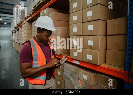 Ethnischer Arbeiter, der durch ein digitales Tablet scrollt, um Pakete zu finden, die zur Auslieferung in der Fabrik geschickt wurden. Stockfoto