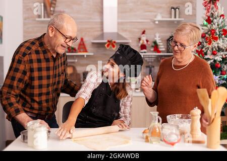 Lächelnde Familie, die in der mit Weihnachten dekorierten kulinarischen Küche am Tisch steht, um die weihnachtsfeiertage zu feiern und die Wintersaison zu genießen. Kind mit Schürze macht Kekse hausgemachten Teig Kochen Lebkuchen Dessert Stockfoto