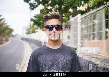 Porträt eines gutaussehenden Jungen im Teenageralter, der eine Sonnenbrille trägt und auf der Straße neben einem Metallzaun steht. Lächelnder selbstbewusster Teenager-Junge in Sonnenbrillen lo Stockfoto