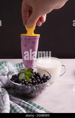 Eine Frauenhand hält auf dunklem Grund hausgemachtes Eis mit Beeren über einen Teller Heidelbeeren. Stockfoto