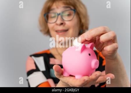 Eine ältere Frau mit Brille legt eine Münze in ein Sparschwein auf weißem Hintergrund Stockfoto