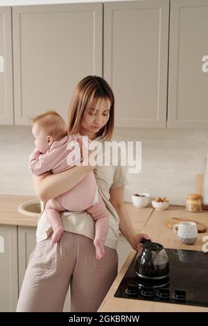 Junge Mutter mit entzückenden Baby Tochter stehen am Elektroherd und nehmen Teekannen Stockfoto