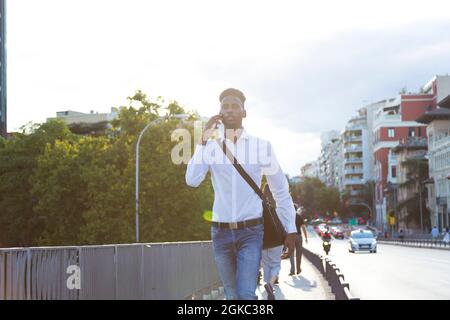 Porträt eines jungen afroamerikanischen Geschäftsmannes mit Aktentasche, die am Telefon spricht und durch die Stadt läuft Stockfoto