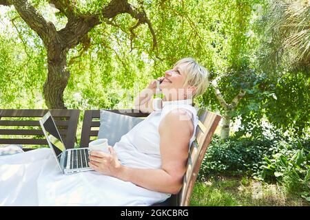 Im Sommer nutzt eine ältere Frau als Freiberuflerin das Smartphone am Telefon im Garten Stockfoto